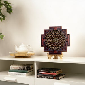 May include: A wooden wall hanging with a geometric design in shades of red, blue, and gold. The design is a Sri Yantra, a sacred symbol in Hinduism. The wall hanging is on a white shelf with books and a teapot.