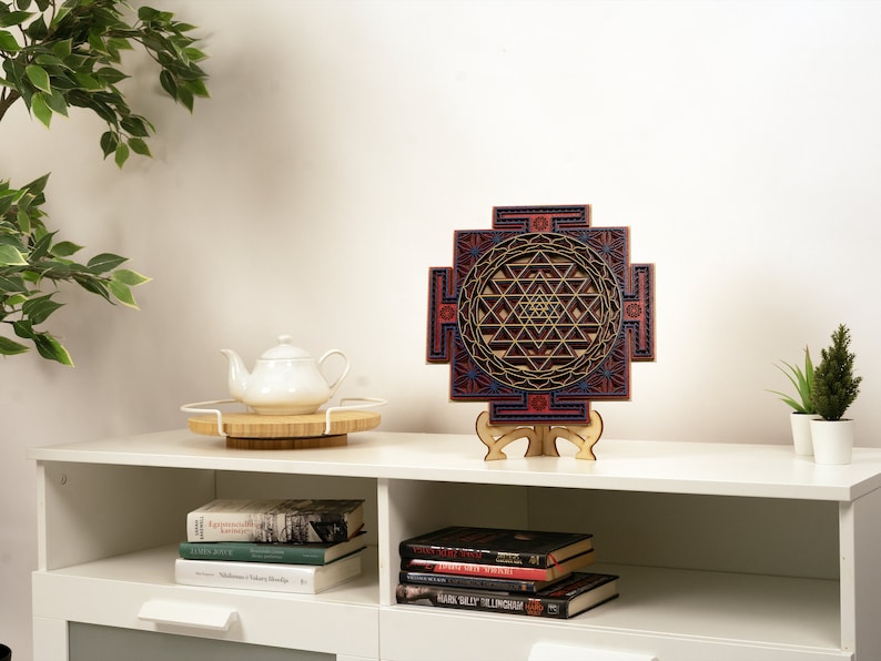 May include: A wooden wall hanging with a geometric design in shades of red, blue, and gold. The design is a Sri Yantra, a sacred symbol in Hinduism. The wall hanging is on a white shelf with books and a teapot.