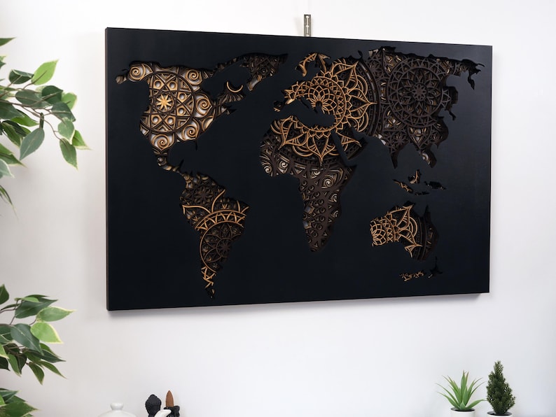 Puede incluir: Un mapa del mundo de madera negra con un dise&ntilde;o 3D que presenta intrincados patrones de mandala dorados. El mapa est&aacute; montado sobre un fondo negro.