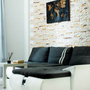 Puede incluir: Un sof&aacute; seccional blanco y gris con un coj&iacute;n de tela negro y una decoraci&oacute;n de pared de mapa del mundo negro y dorado en una pared de ladrillo blanco.