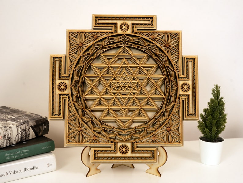 May include: A wooden wall hanging with a geometric design, featuring a Sri Yantra, a sacred symbol in Hinduism. The wall hanging is on a wooden stand and has a light brown color.