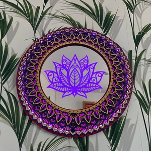 May include: A round wooden mirror with a purple mandala design and a purple lotus flower in the center. The mirror is surrounded by a decorative frame with a red and gold border.