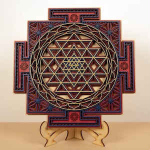 May include: A wooden wall hanging with a geometric design in shades of red, blue, and gold. The design is a Sri Yantra, a sacred symbol in Hinduism.