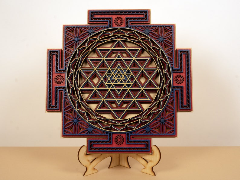 May include: A wooden wall hanging with a geometric design in shades of red, blue, and gold. The design is a Sri Yantra, a sacred symbol in Hinduism.