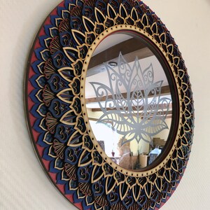 May include: Round wooden mirror with a decorative mandala design in shades of red, blue, and brown. The mirror has a clear glass surface with a cutout of a lotus flower.