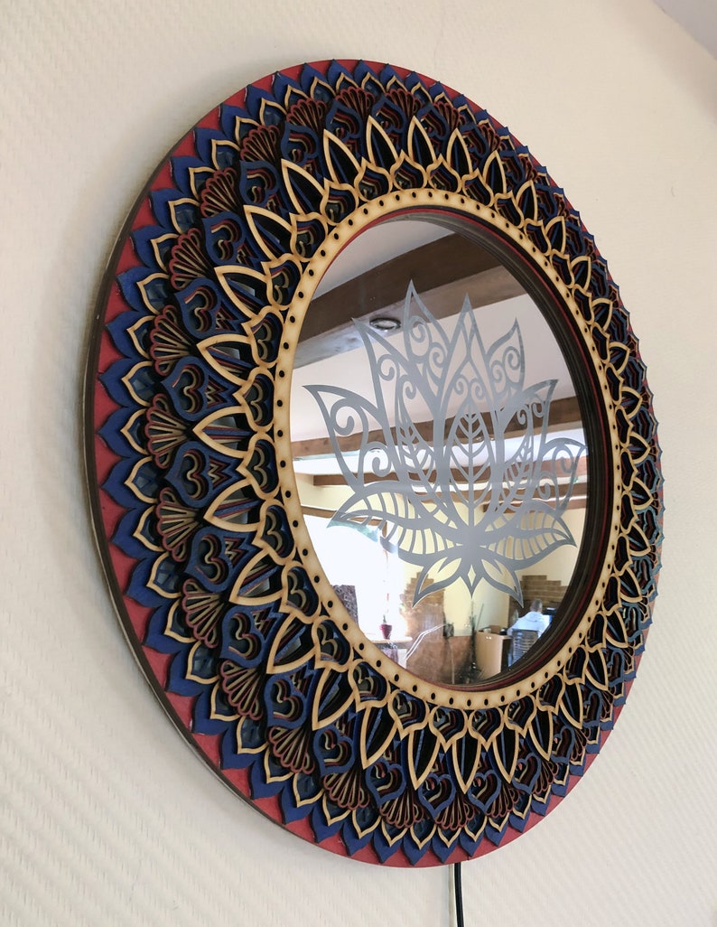 May include: Round wooden mirror with a decorative mandala design in shades of red, blue, and brown. The mirror has a clear glass surface with a cutout of a lotus flower.