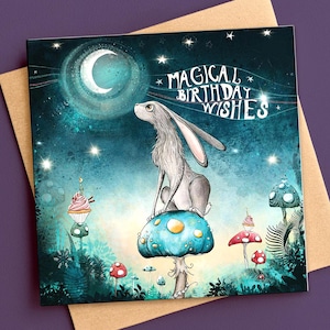 May include: A greeting card with a whimsical illustration. A gray hare sits atop a blue mushroom, gazing at a crescent moon. The card features the text "MAGICAL BIRTHDAY WISHES" and is set against a starry, teal background. Includes a brown envelope.