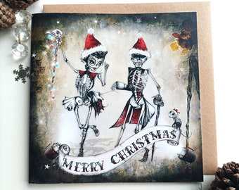 May include: A square greeting card featuring an illustration of two skeletons wearing red and white Santa hats. The skeletons are dancing above a banner that reads MERRY CHRISTMAS. A small cat sits near the banner, and the background is dark.