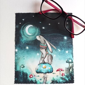 Può includere: Un panno per la pulizia in microfibra nero con un'illustrazione fantasiosa di un coniglio seduto su un fungo sotto una luna crescente e stelle. Il panno è circondato da uno sfondo bianco.