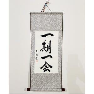 Puede incluir: Un rollo japonés tradicional con caligrafía en tinta negra sobre papel blanco. El rollo está enmarcado por una tela gris estampada y tiene varillas de madera en la parte superior e inferior. El texto dice "Ichi-go ichi-e".