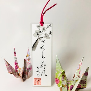 Peut inclure: Un marque-page avec un motif de fleurs de cerisier et de la calligraphie japonaise, avec un cordon rouge. Deux grues en origami aux motifs floraux encadrent le marque-page. Le texte "Wind blowing cherry blossoms" est en haut.