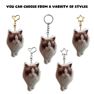 May include: Five plush cat keychains with different colored metal charms. The charms are a heart, a circle, a star, a heart, and a star. The keychains are all white with a brown and white cat face printed on them.