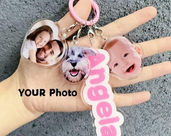 May include: A collection of personalized keychains featuring custom photos. The keychains include a heart-shaped one with a family photo, a dog's face, a baby's face, and a pink name tag that says "Angela". They are attached to a pink keyring.