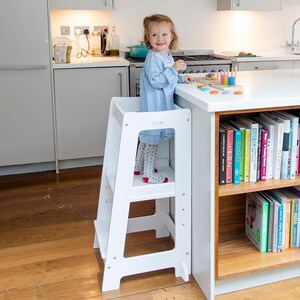 May include: A white wooden kitchen step stool with two steps and a platform. The stool has the word "ELEVATE" printed on the side. A child is standing on the platform, near a white countertop with colourful toys.