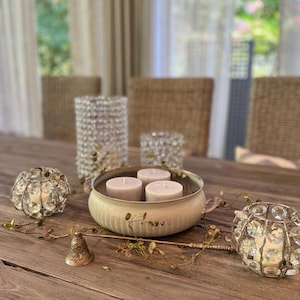 May include: A rustic wooden table with a white ceramic bowl filled with three white pillar candles. The bowl has a decorative handle and is surrounded by crystal candle holders. A brass candle snuffer is on the table.