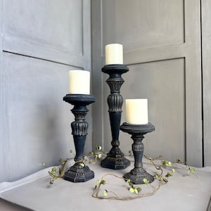 May include: Three black decorative candle holders with white candles. The holders have ornate detailing and are arranged on a gray surface. A garland of green leaves and small berries is draped across the surface.