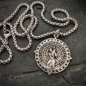 Op de afbeelding: Een zilveren ketting met een ronde hanger met een afbeelding van Sint Christoffel die een kind draagt. De hanger is gegraveerd met "St. Christopher Protect Us" en een kompasroos.