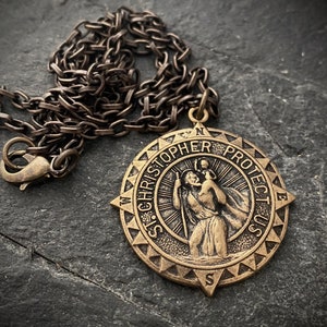 Op de afbeelding: Een bronzen ketting met een hanger in de vorm van een kompas met de tekst "St. Christopher Protect Us". De hanger toont een figuur van Sint Christoffel die een kind op zijn schouders draagt.