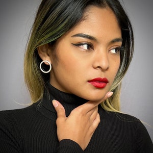 Könnte beinhalten: Eine Frau mit langen dunklen Haaren trägt ein schwarzes Rollkragen-Shirt und einen silbernen Creolen-Ohrring. Sie schaut in die Kamera und hat die Hand am Kinn.