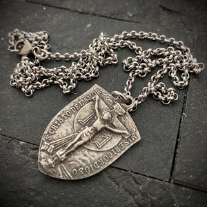 May include: A silver-toned necklace featuring a pendant with a religious motif. The pendant depicts a crucified figure and is inscribed with text. The chain is a linked design, and the piece rests on a dark, textured surface.
