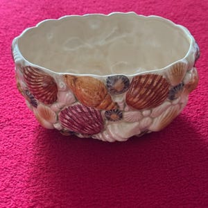 May include: Cream ceramic bowl with a scalloped edge, decorated with assorted seashells in brown, pink, and cream hues. The bowl features a textured exterior and a smooth interior, suitable for decorative purposes.