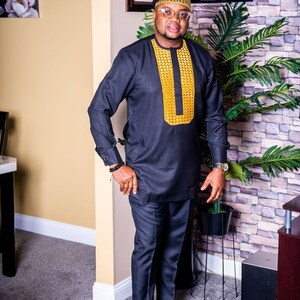 May include: A man in a black outfit with gold embroidery on the shirt. He wears a matching hat, glasses, and black shoes with gold details. The background features framed pictures and a potted plant. The outfit is a traditional style.
