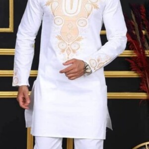 May include: A white tunic and trousers outfit with gold embroidery around the neckline and cuffs. The person is wearing black sunglasses and shoes. The style is a traditional design.