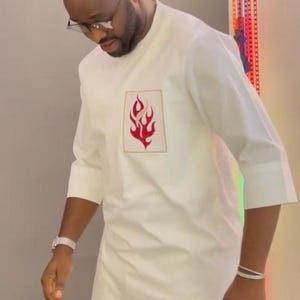 May include: A white tunic featuring a red and gold flame emblem. The garment has three-quarter length sleeves and a refined, understated design. Suitable for formal or semi-formal events.