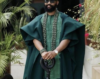 May include: A man in a dark green traditional outfit, including a hat, tunic, and cape. He wears sunglasses and holds a black, ornate cane. The outfit features intricate embroidery and is accessorized with a beaded necklace and bracelet. The setting is outdoors with greenery.