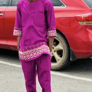 May include: A magenta two-piece outfit, comprising a long-sleeved shirt and matching trousers. The shirt has decorative trim on the sleeves and waist. The person is wearing brown sandals. A red car is in the background.