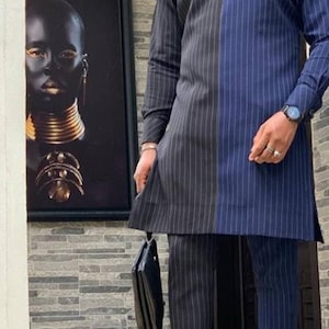 May include: A two-tone, long-sleeved suit with vertical pinstripes in navy blue and black. The outfit is paired with matching trousers and patterned loafers. A black handbag is held in one hand. A framed portrait hangs on the wall behind the person.