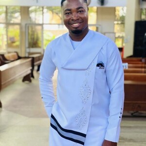 May include: A white tunic with long sleeves, a draped neckline, and decorative detailing. The tunic has a black and grey stripe and is worn with white trousers. The person is smiling in a bright room with wooden benches.