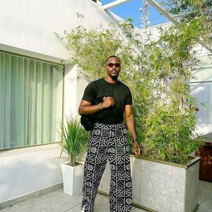 May include: A person wearing a black t-shirt and black and white patterned trousers. The trousers have a geometric design. The person is wearing white trainers and a black bag. The background includes greenery and a building.