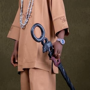 May include: A person in a tan traditional outfit, including a hat, tunic, and trousers. The outfit is accessorized with a beaded necklace and a decorative staff. The staff is black with a carved animal figure at the top.