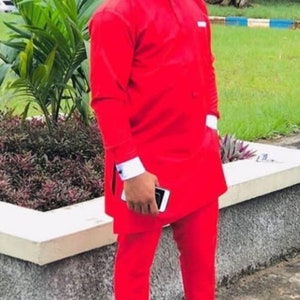May include: A man in a vibrant red suit, featuring a mandarin collar shirt and trousers. The outfit includes white cuffs and black loafers with gold detailing. The background shows greenery and a concrete structure.