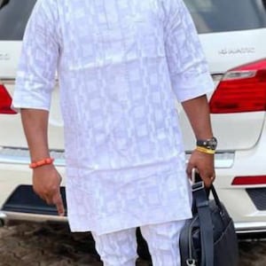 May include: A man in a white patterned outfit, red cap, and sunglasses stands in front of a white SUV. He carries a black bag and wears black loafers with gold accents. The outfit includes a short-sleeved shirt and matching trousers.