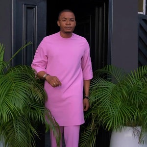 May include: A pink two-piece suit, comprising a tunic with decorative sleeve slits and matching trousers. The tunic has a round neck. The suit is worn with black shoes. The logo "Am Diddy, A style for every story..." is visible.