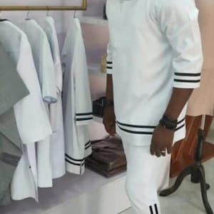 May include: A white two-piece outfit with black stripes on the sleeves and trousers. The outfit is worn with black shoes. Several white garments hang on a rail in the background.