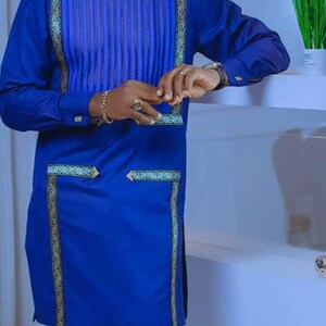 May include: A blue tunic and trousers ensemble. The tunic has vertical pleats on the chest and gold decorative trim. The outfit is a solid blue colour. The trousers are a matching blue.