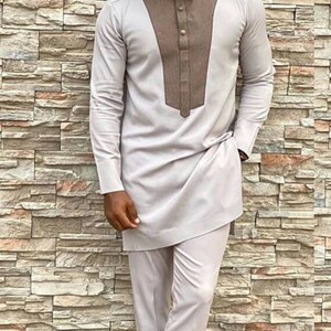 May include: A light grey two-piece suit with a brown panel on the shirt's chest. The long-sleeved shirt and trousers are matching. The outfit is paired with brown leather loafers. The background is a stone wall.