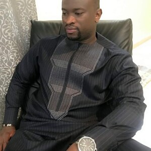 May include: A dark navy blue, long-sleeved, striped shirt and matching trousers. The shirt has a geometric design on the chest. A silver wristwatch with a black dial is visible on the wrist. The outfit is formal and tailored.