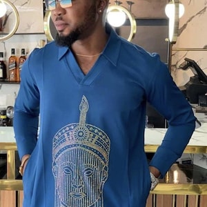May include: A long-sleeved, blue tunic with a V-neck and a decorative face design on the front. The design is made of small, sparkling embellishments. The tunic has pockets and is worn with sunglasses.