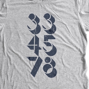 Może przedstawiać: Szary T-shirt z graficznym wzorem przedstawiającym cyfry 3, 3, 4, 5, 7 i 8 ułożone pionowo. Cyfry są w pogrubionym, blokowym kroju i są nadrukowane ciemnoniebieskim kolorem.