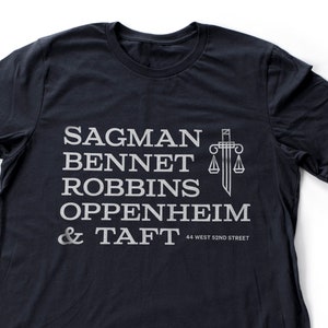 Könnte beinhalten: Ein dunkelblaues T-Shirt mit einem weißen Grafikdesign, das den Text "Sagman Bennet Robbins Oppenheim & Taft" und ein stilisiertes Bild eines Schwertes und einer Waage zeigt. Der Text "44 West 52nd Street" ist ebenfalls im Design enthalten.