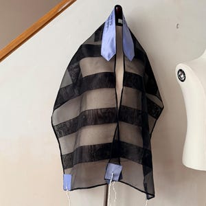 May include: A black and sheer striped prayer shawl with light blue accents. The shawl is hanging on a black hanger. The shawl has a light blue collar and light blue squares at the bottom with white tassels.