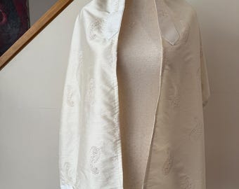 May include: Cream-coloured prayer shawl with embroidered paisley patterns. The shawl is draped over a mannequin, showcasing its rectangular shape and delicate fringe. The fabric appears to be silk or a similar material.