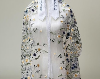May include: A white sheer tallit featuring an intricate floral embroidery pattern in shades of yellow, blue, and green. The garment includes a white atarah with Hebrew text embroidered in blue, finished with a delicate lace trim and white fringe.