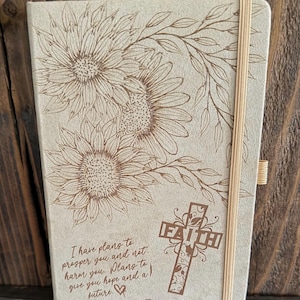 May include: A light beige notebook with a brown laser-engraved design of three sunflowers and leafy vines. The cover features a cross with the word FAITH and the text I have plans to prosper you and not harm you. Plans to give you hope and a future. A tan elastic band secures the notebook.