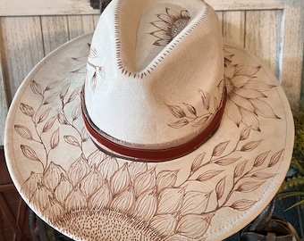 May include: A white cowboy hat with a brown leather band and a sunflower design painted on the crown and brim.