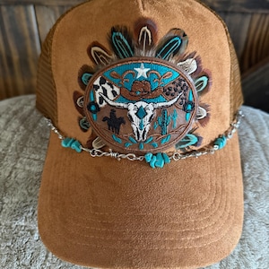May include: A tan suede trucker hat featuring a central oval patch with embroidered designs of a longhorn skull, a cowboy hat, a cactus, and a rider on horseback. The patch is surrounded by brown and turquoise feathers and accented by a silver chain with turquoise stones.